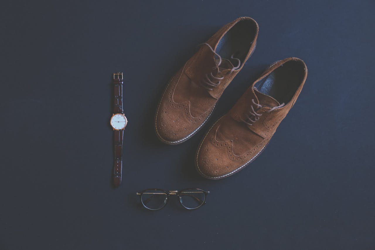 why-choose-us Flat lay of brown leather shoes, eyeglasses, and wristwatch on a dark background.